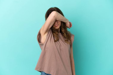 Orta yaşlı beyaz kadın, mavi arka planda izole edilmiş, gözleri elle kapatılmış.