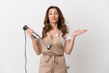 Orta yaşlı beyaz kadın el blenderi kullanıyor. Beyaz arka planda izole edilmiş, şok olmuş bir yüz ifadesi var.