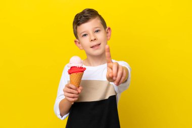 Kornet dondurmalı küçük beyaz çocuk sarı arka planda izole edilmiş ve parmağını kaldırıyor.