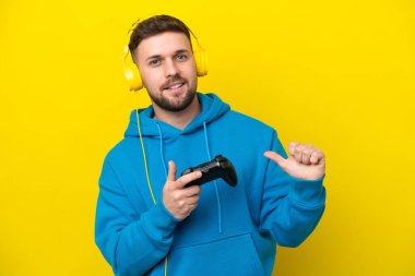 Sarı arka planda izole edilmiş, gururlu ve kendini beğenmiş bir bilgisayar oyunu kontrolörüyle oynayan beyaz bir genç.
