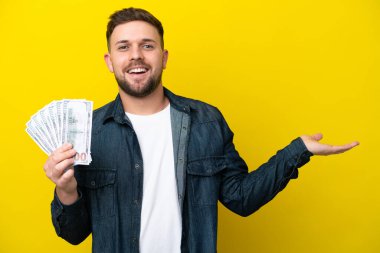 Kafkasyalı genç adam, sarı arka planda izole edilmiş bir sürü para alıyor. Davet etmek için ellerini yana uzatıyor.