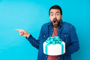 Büyük pastası olan genç ve yakışıklı bir adam. Mavi arka planda. Şaşırmış ve parmağıyla işaret ediyor.