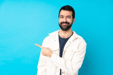 Beyaz kadife ceketli, mavi arka planlı genç yakışıklı bir adam bir ürünü sunmak için yan tarafı işaret ediyor.