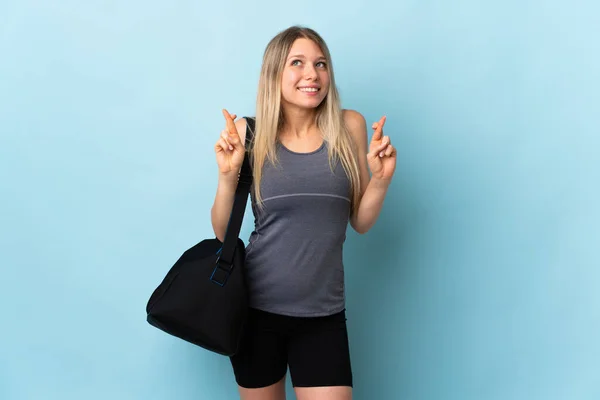 Young sport woman with sport bag isolated on blue background with fingers crossing and wishing the best