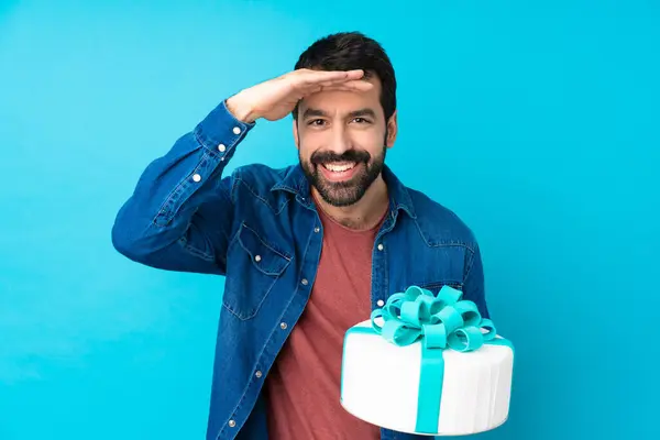 Young handsome man with a big cake over isolated blue background looking far away with hand to look something