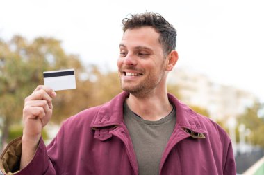 Açık havada elinde mutlu bir ifadeyle kredi kartı tutan genç beyaz bir adam.