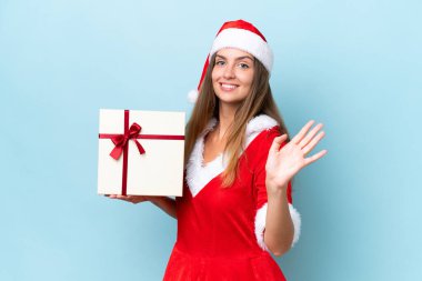 Anne Noel gibi giyinmiş genç beyaz kadın elinde hediyeyle mavi arka planda izole edilmiş mutlu bir ifadeyle selam veriyor.