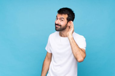 Mavi arka planda sakallı genç adam kulağına elini sürerek bir şeyler dinliyor.