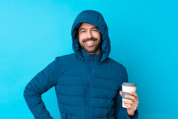 Man wearing winter jacket and holding a takeaway coffee over isolated blue background posing with arms at hip and smiling