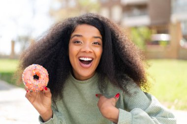 Genç Afrikalı Amerikalı bir kadın açık havada yüzünde sürpriz bir ifadeyle donut tutuyor.