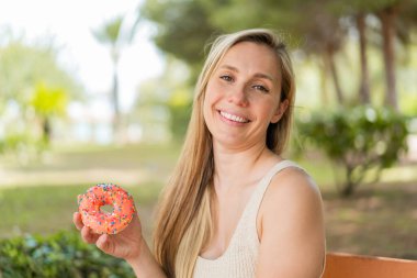 Sarışın genç bir kadın açık havada donut tutuyor ve gülümsüyor.