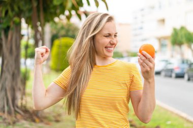 Açık havada elinde portakal tutan genç sarışın kadın zaferi kutluyor.
