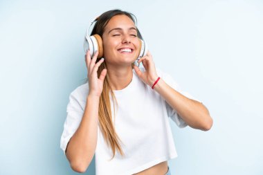 Genç beyaz kadın mavi arka planda izole edilmiş müzik dinliyor ve şarkı söylüyor.