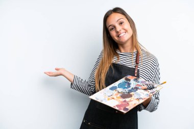 Beyaz arka planda izole edilmiş bir palet tutan beyaz tenli genç bir kadın davet etmek için ellerini yana uzatıyor.