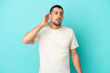 Genç beyaz adam mavi arka planda izole edilmiş bir şekilde elini kulağına koyarak bir şeyler dinliyor.
