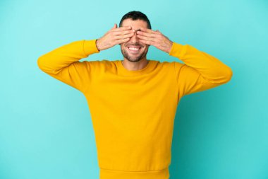 Genç yakışıklı beyaz adam mavi arka planda izole edilmiş gözleri elleriyle kaplıyor ve gülümsüyor.
