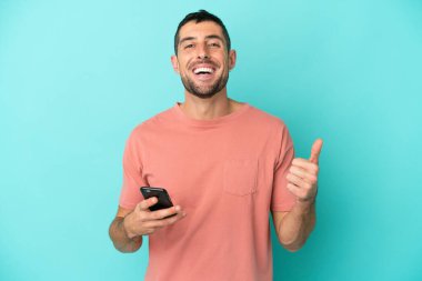 Young handsome caucasian man isolated on blue background using mobile phone while doing thumbs up