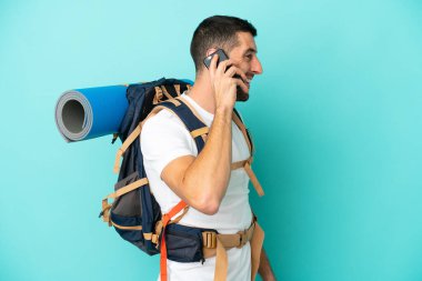 Mavi arka planda izole edilmiş büyük bir sırt çantası olan genç bir beyaz adam cep telefonuyla sohbet ediyor.