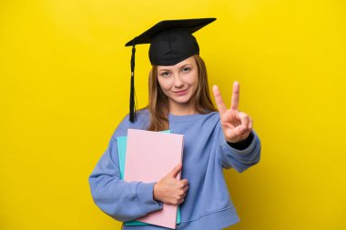 Sarı arka planda izole edilmiş genç Rus kadın gülümsüyor ve zafer işareti gösteriyor