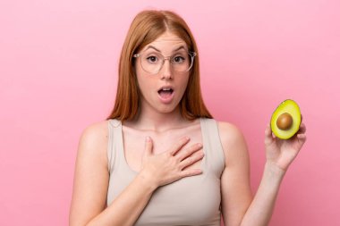 Elinde avakado tutan genç kızıl saçlı kadın pembe arka planda izole edilmiş, şaşırmış ve şaşırmış bir şekilde sağa bakıyordu.