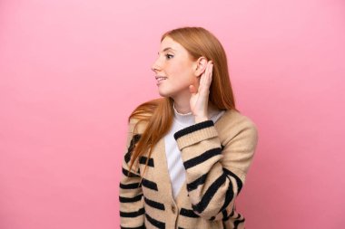 Genç kızıl saçlı kadın pembe arka planda izole edilmiş. Kulağına elini sürerek bir şeyler dinliyor.