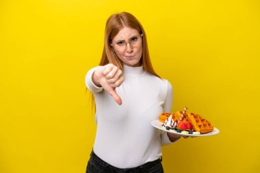 Sarı arka planda gözleme tutan genç kızıl saçlı kadın olumsuz bir ifadeyle baş parmağını aşağı gösteriyor.