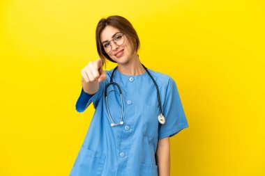 Doktor kadın, sarı arka planda izole edilmiş. Parmağını kendine güvenen bir ifadeyle işaret ediyor.