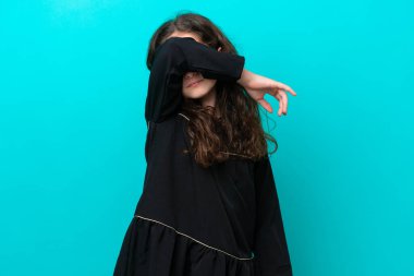 Mavi arka planda izole edilmiş küçük bir kız gözlerini elleriyle kapatıyor.