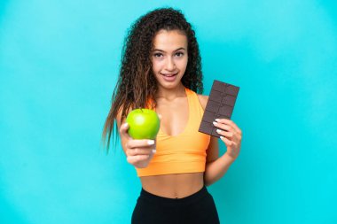 Genç bir Arap kadın mavi arka planda izole edilmiş. Bir elinde çikolata tableti, diğerinde elma alıyor.