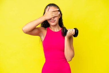 Genç sporcu kadın halter yapıyor. Sarı arka planda izole edilmiş. Gözlerini elleriyle kapatıyor. Bir şey görmek istemiyorum.