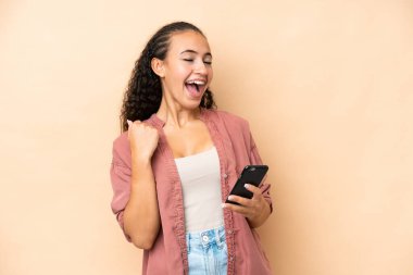 Galibiyet pozisyonunda telefonla bej arka planda izole edilmiş genç bir kadın