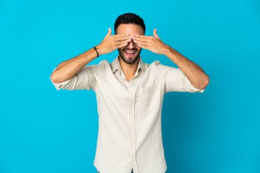 Mavi arka planda izole edilmiş genç beyaz bir adam elleriyle gözlerini kapatıyor ve gülümsüyor.
