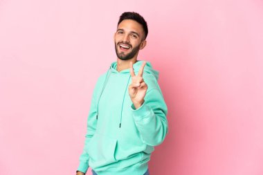 Pembe arka planda izole edilmiş genç beyaz adam gülümsüyor ve zafer işareti gösteriyor.