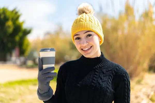 Young moroccan girl wearing winter muffs while holding a coffee at outdoors smiling a lot