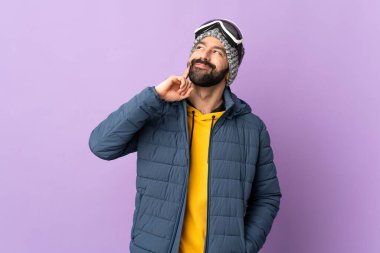 Kar kaykayı gözlüklü kayakçı mor arka planda yukarı bakarken bir fikir düşünüyor.