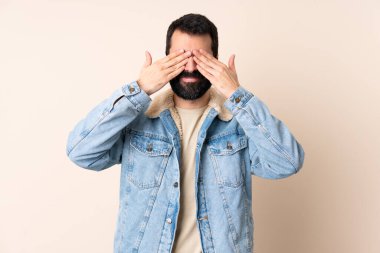 İzole edilmiş arka planda sakallı, gözleri elleriyle kaplayan ve gülümseyen beyaz bir adam.