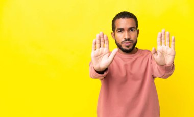 Sarı arka planda izole edilmiş genç latin adam dur hareketi yapıyor ve hayal kırıklığına uğruyor.