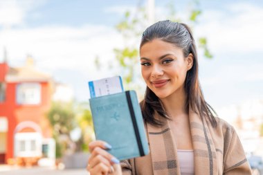 Genç ve güzel bir kadın elinde pasaportuyla dışarıda mutlu bir ifadeyle.