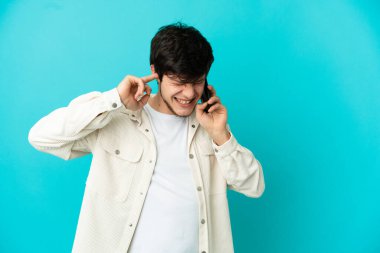 Mavi arka planda izole edilmiş cep telefonu kullanan genç Rus adam hayal kırıklığına uğramış ve kulaklarını kapatmış.