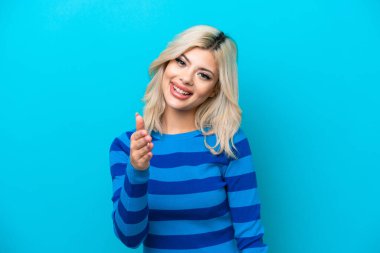 Mavi arka planda izole edilmiş genç bir Rus kadın iyi bir anlaşma için el sıkışıyor.