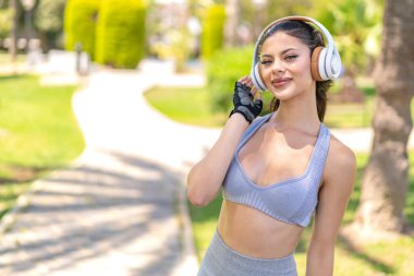 Açık havada genç ve güzel bir kadın spor giysileri giyiyor ve kulaklıkla müzik dinliyor.