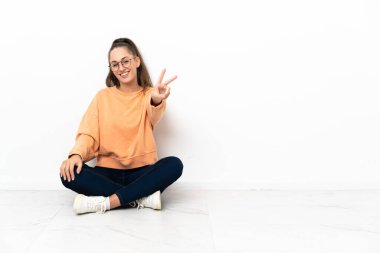 Yerde oturmuş gülümseyen ve zafer işareti gösteren genç bir kadın.