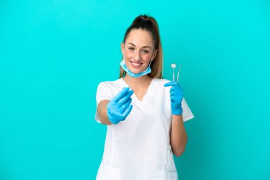 Elinde mavi arka planda izole edilmiş aletler tutan beyaz dişçi kadın para hareketi yapıyor.