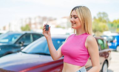 Genç, güzel sarışın kadın arabanın anahtarlarını açık havada tutarken mutlu bir ifadeyle.