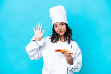 Mavi arka planda sashimi tutan genç İspanyol aşçı kadın parmaklarıyla beşi sayıyor.