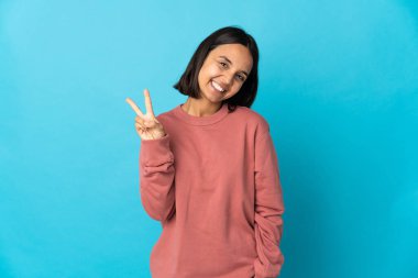 Mavi arka planda izole edilmiş genç Latin kadın gülümsüyor ve zafer işareti gösteriyor.