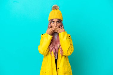 Pembe saçlı, yağmura dayanıklı bir ceket giyen genç bir kadın. Mavi arka planda izole edilmiş, ağzı elleriyle kapatıyor.