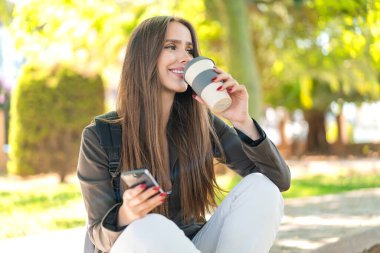 Dışarıda cep telefonu kullanan ve elinde kahve tutan genç bir kadın.