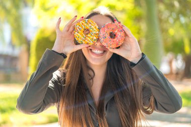 Açık havada genç bir kadın gözlerinde donut tutuyor.