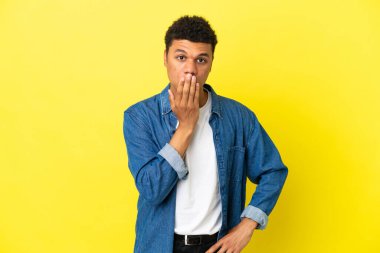Young African American man isolated on yellow background covering mouth with hand